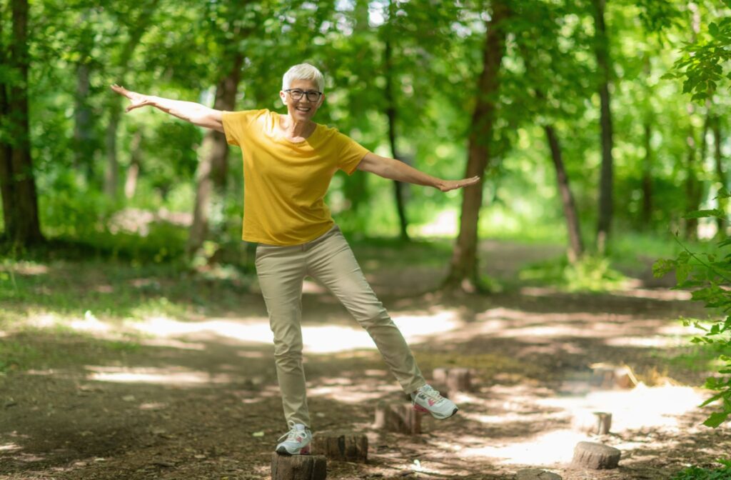 A smiling senior does a single-leg stand on a stump outside.