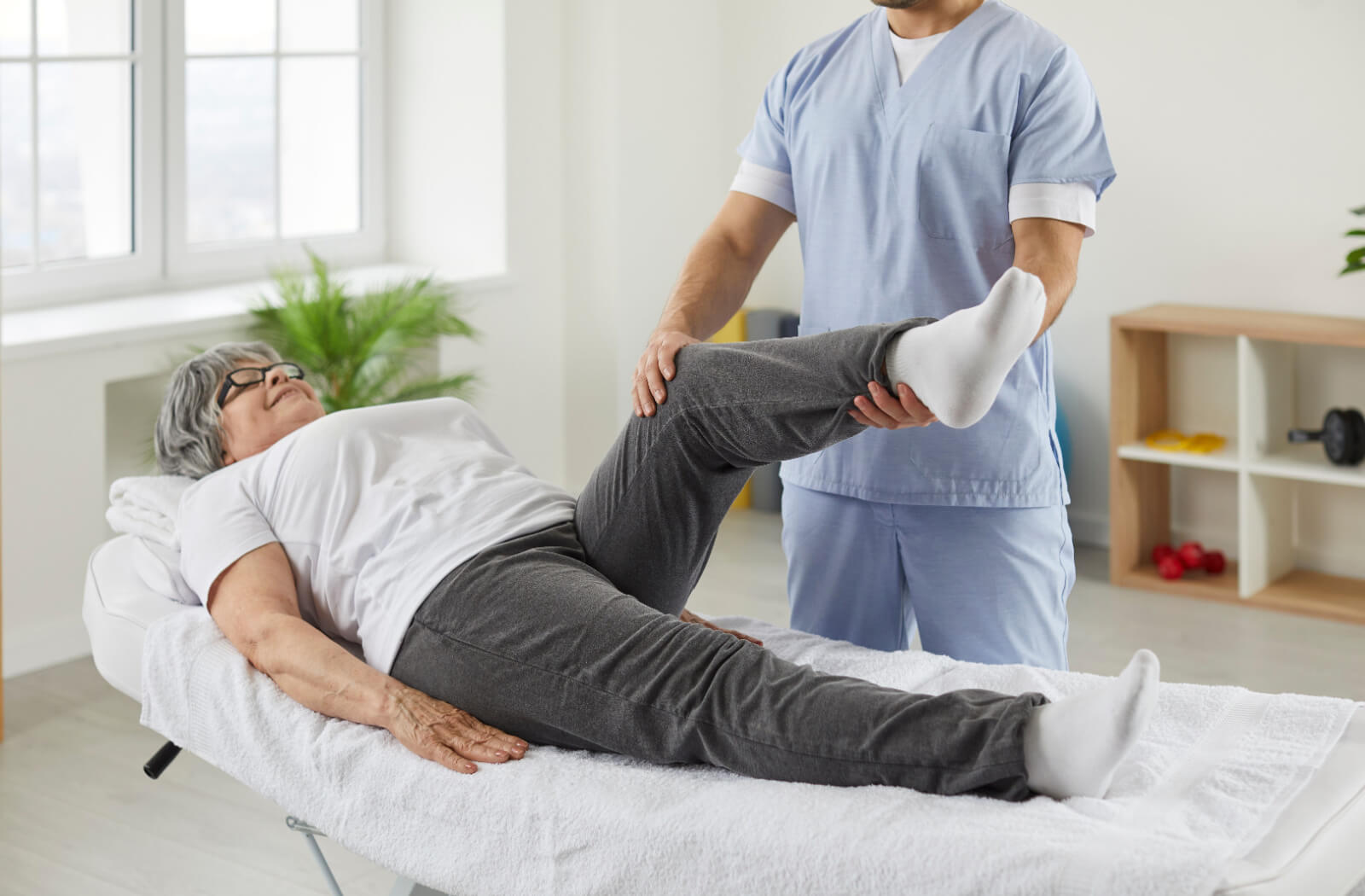 An older adult lying on a table while a physiotherapist helps them stretch their left leg to lower joint discomfort.
