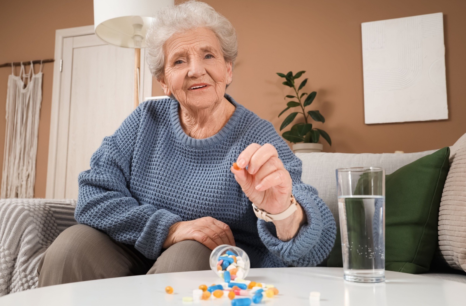 A senior holds a multivitamin pill above a spilled pill container next to a glass of water on a table.