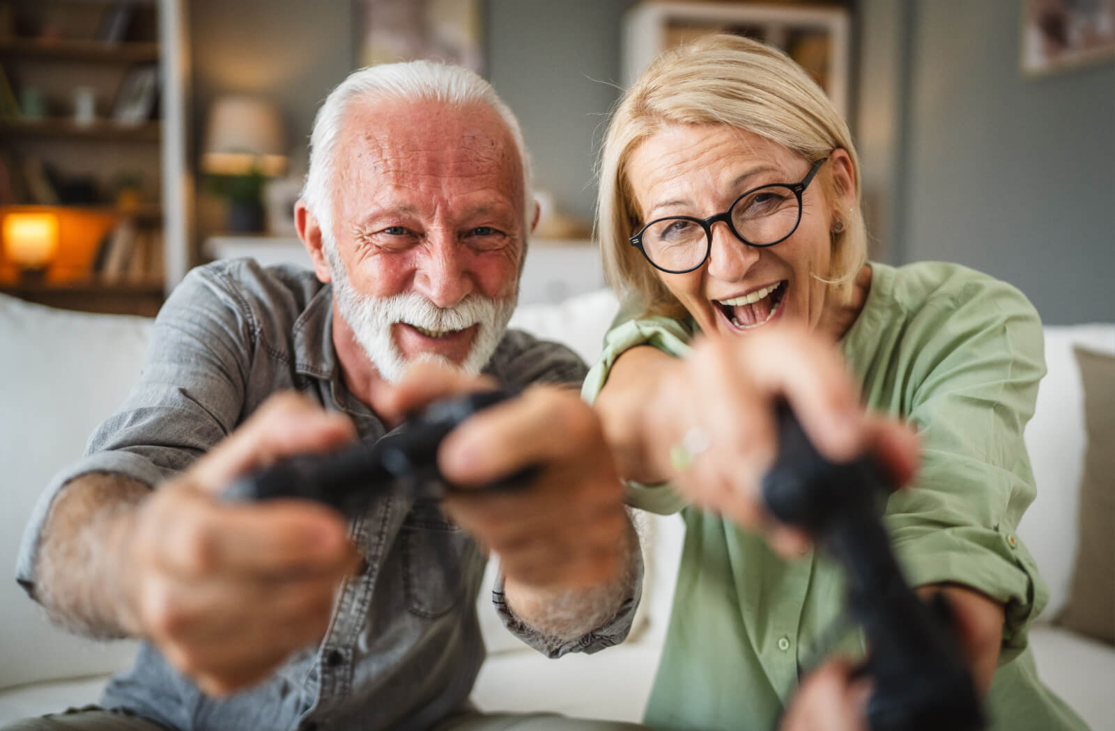 A close-up of a smiling older couple sitting on their couch, holding up video game controllers for their new unusual hobby.