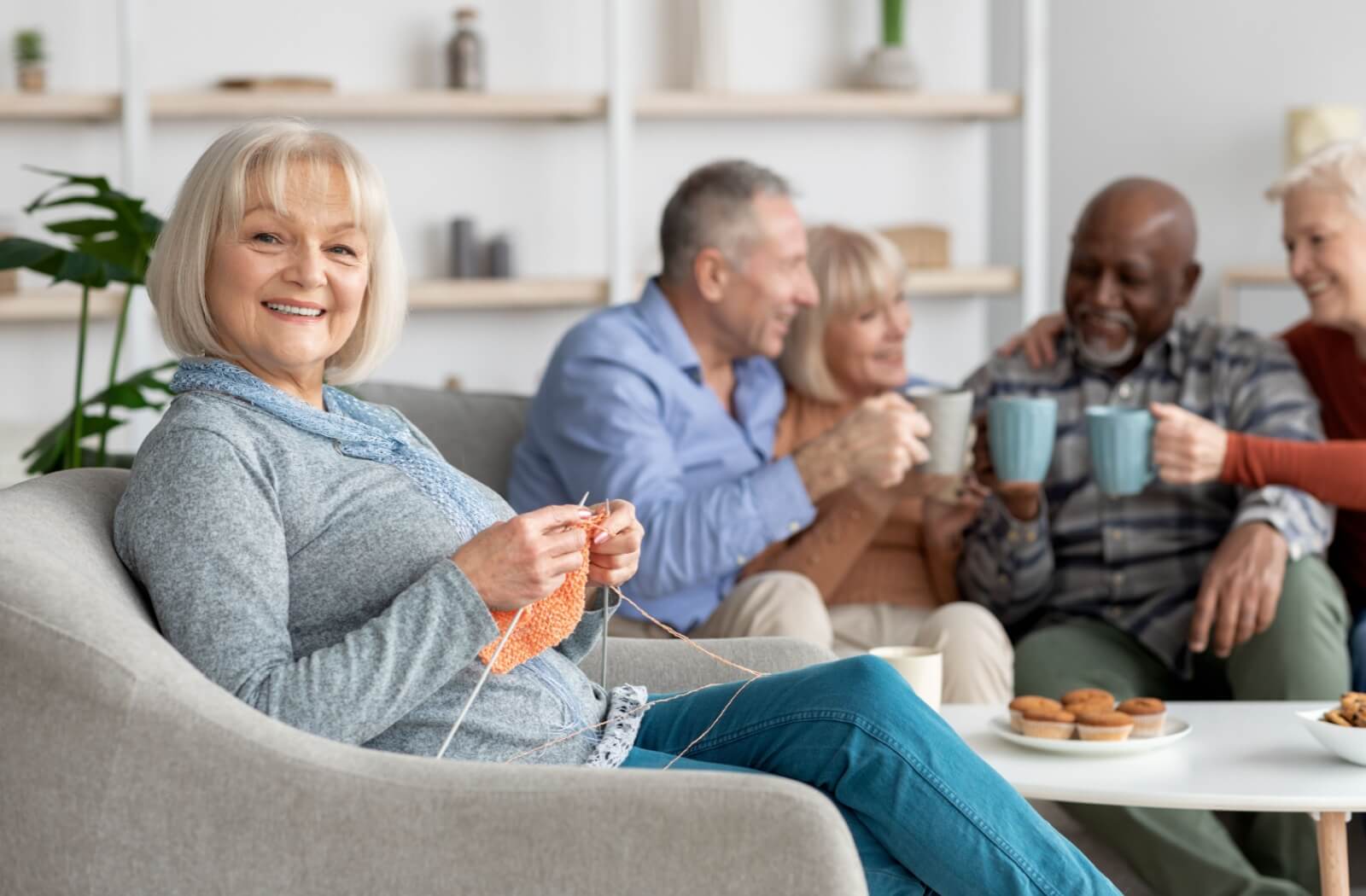 An older woman sitting in a chair smiling and knitting while a group of seniors hold coffee mugs and smile in the background