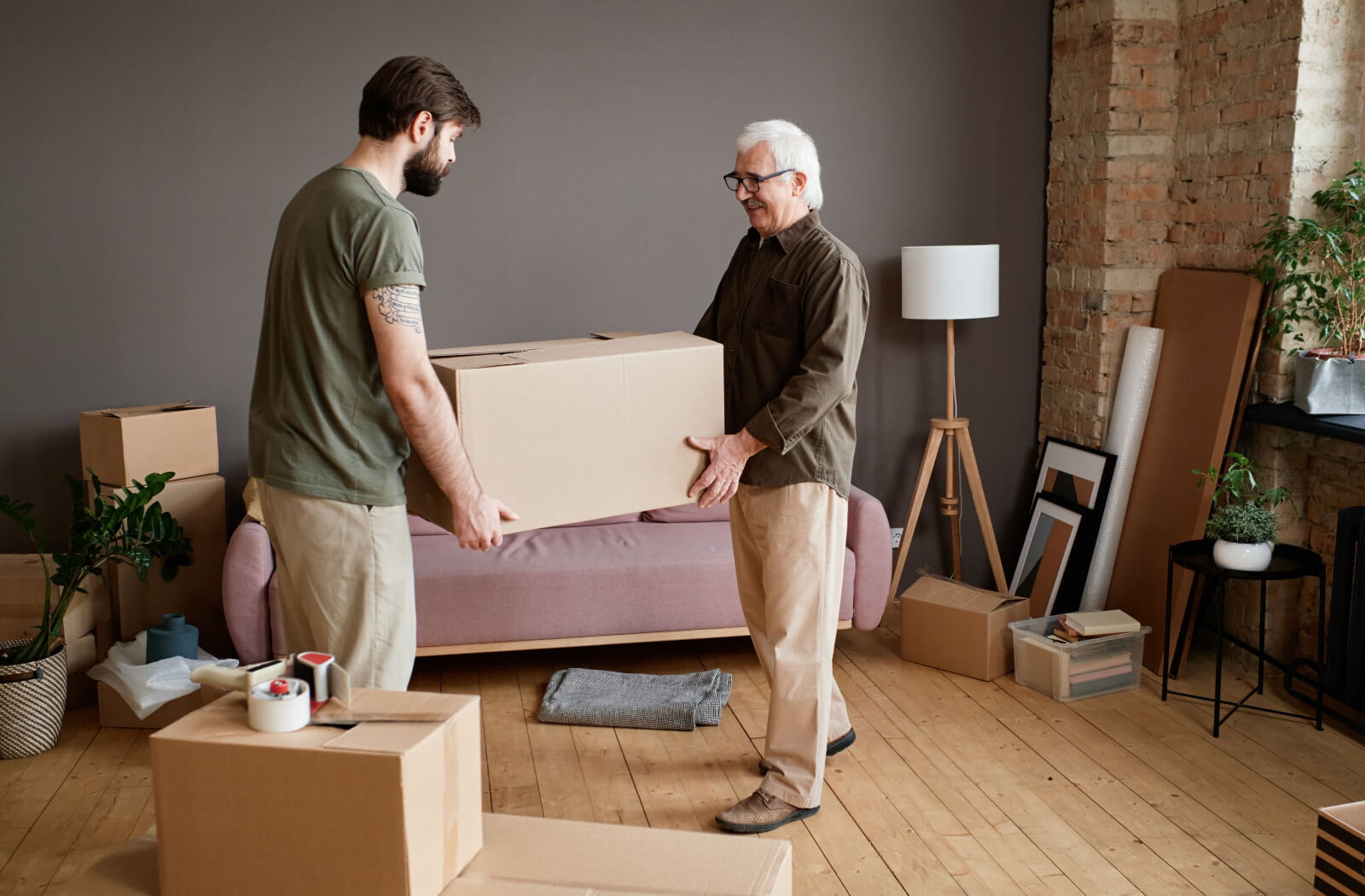 A young man helping a senior man carry a card board box.
