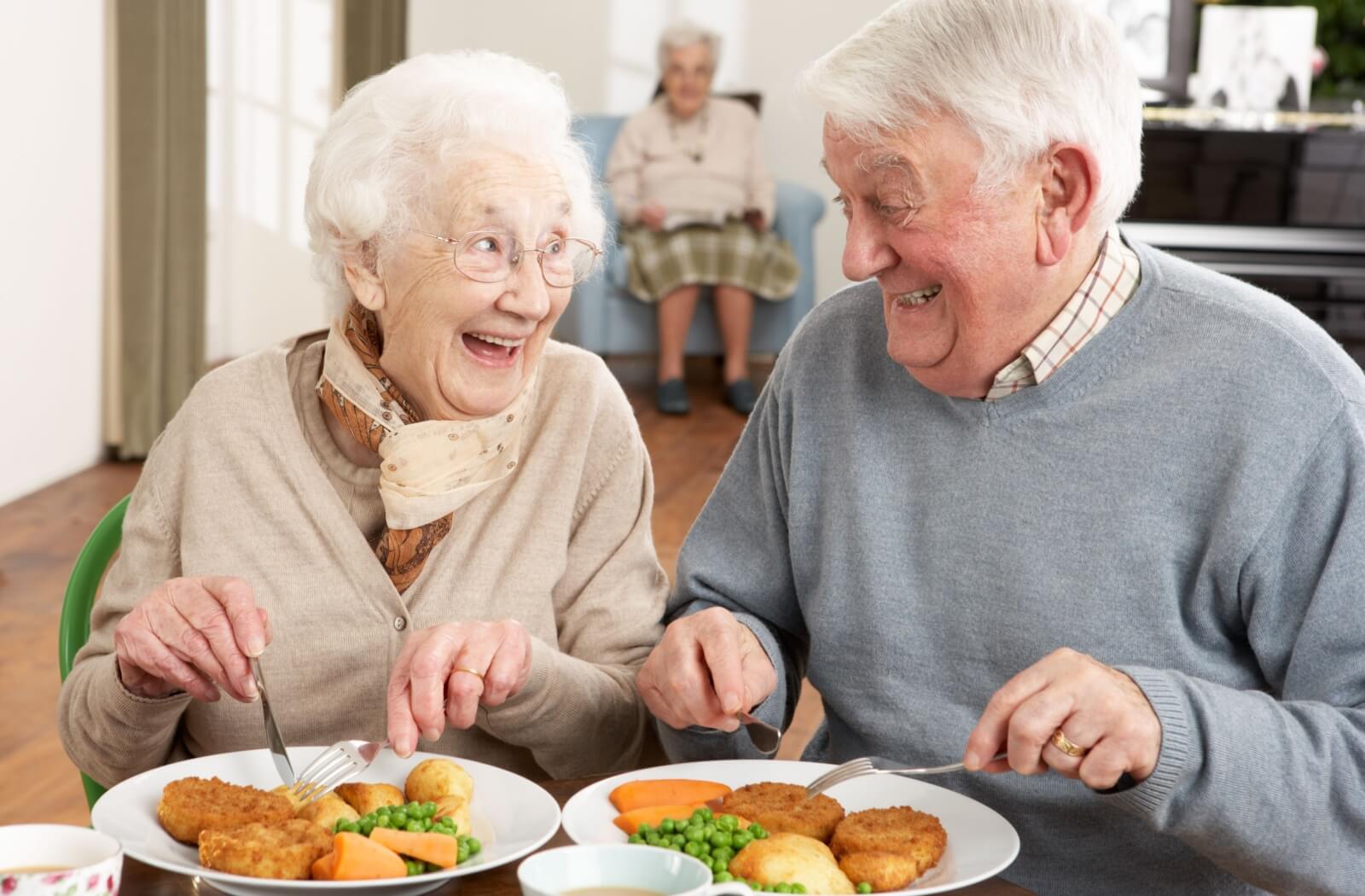 A happy senior couple enjoys a meal in the communal dining area of their senior living community.