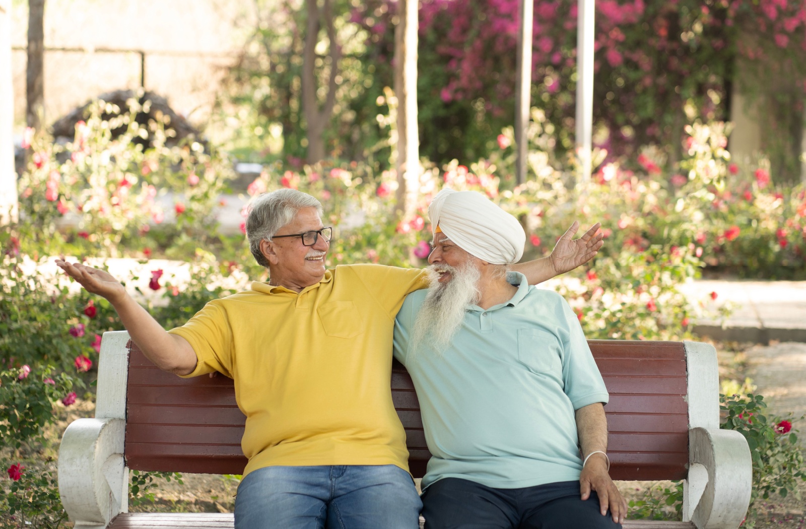 Two happy, close senior friends sit on a park bench together, arms over each other’s shoulders, enjoying the company
