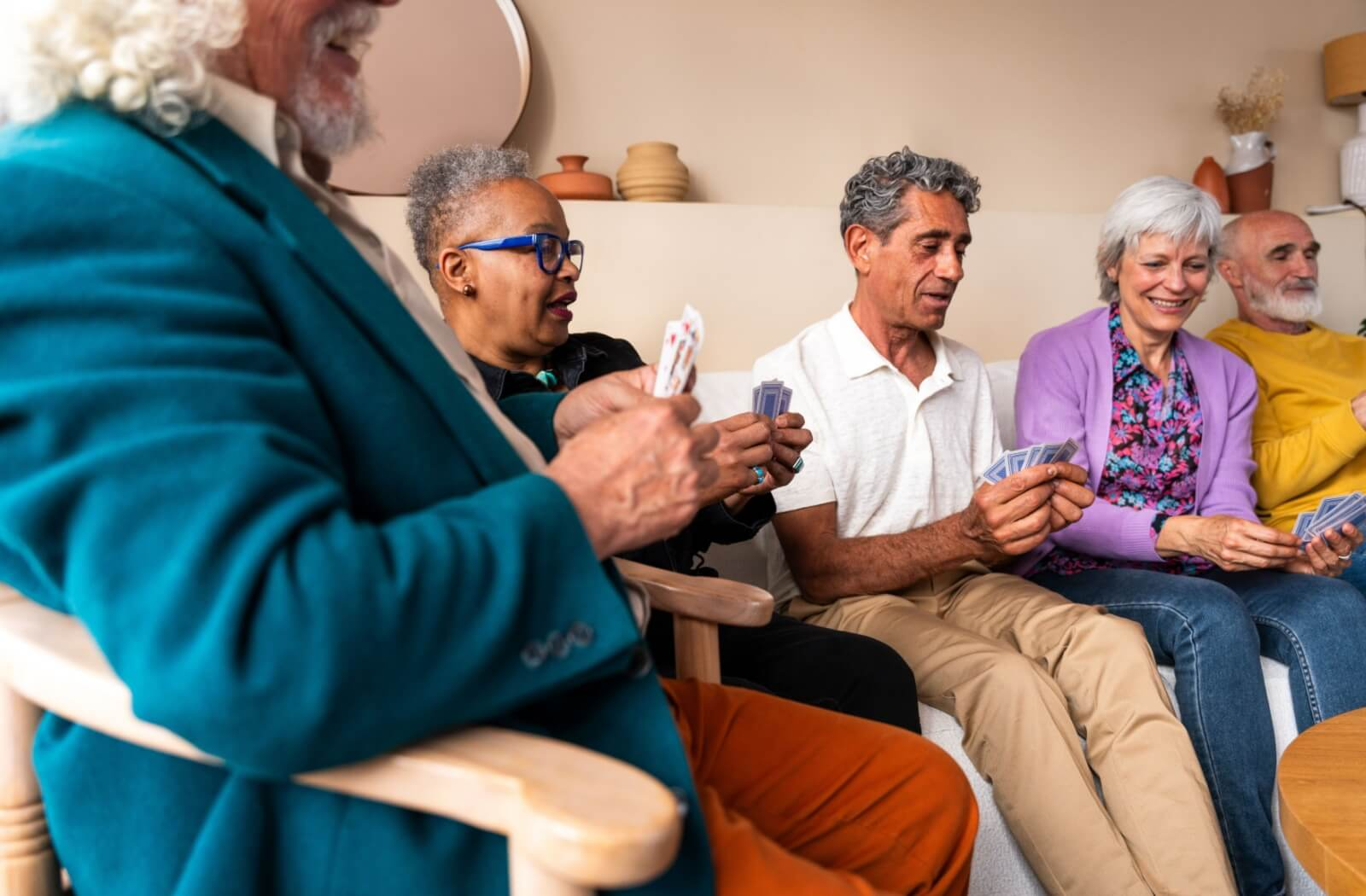 A group of senior friends sit on different pieces of furniture surrounding a circular table to play a big game of cards