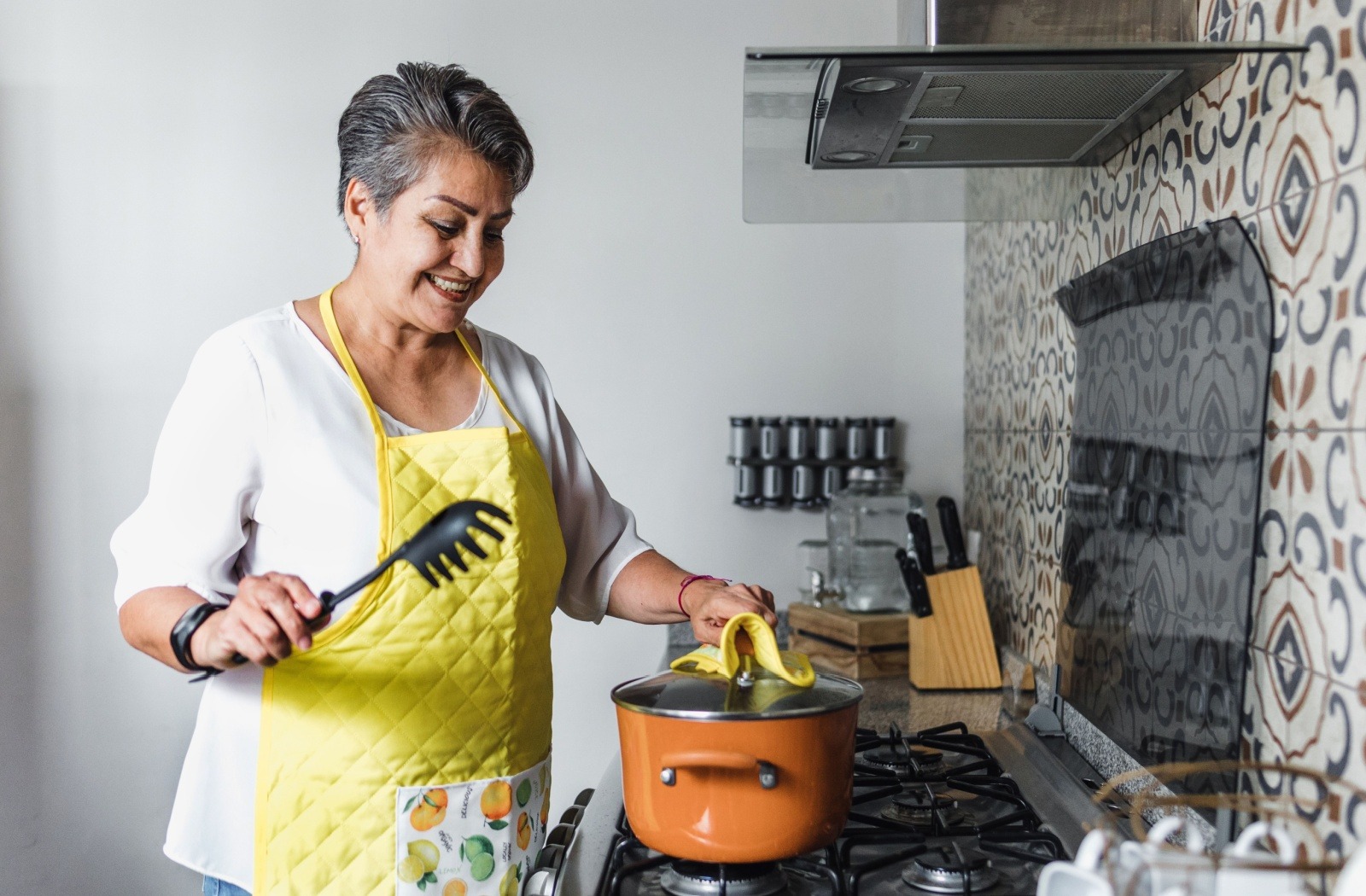 A senior in a yellow apron stands over the stove, smiling while cooking a meal in a bright kitchen setting.
