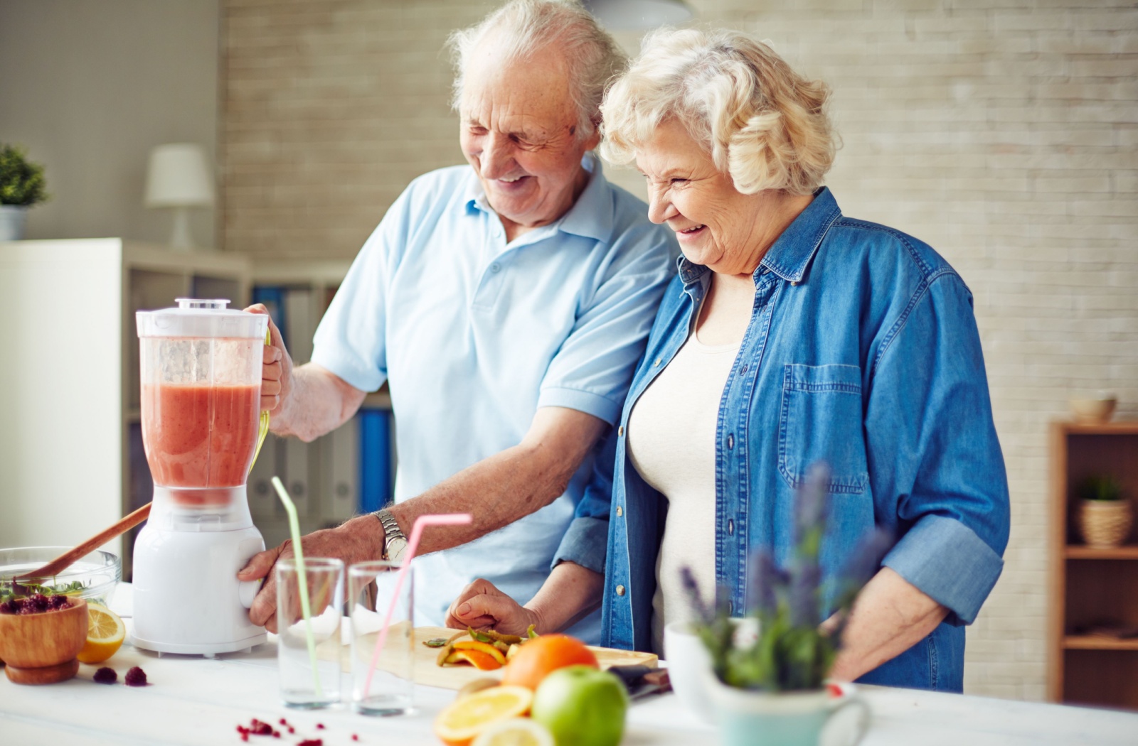 An older couple in senior living stands in their kitchen and makes a smoothie while smiling